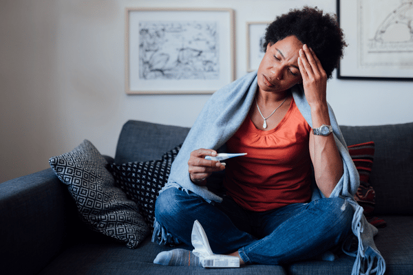 woman sitting on the couch holding her head in one hand and a thermometer in the other. She appears to be sick from some sort of upper respiratory tract infection.