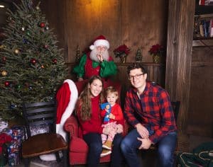 A young couple sit in a red chair with their toddler daughter. Santa stands behind them with his finger up to his lips so no one tells the little girl he is there.