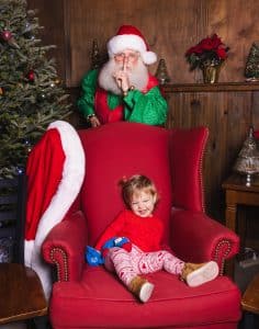 Smiling toddler sits on a chair, oblivious that Santa is behind her
