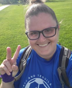 Woman wearing glasses, a backpack and blue soccer jersey, smiling and holding up peace sign with bandaged hand.