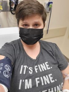 woman in hospital bed, wearing gray shirt reading "It's fine" with blood pressure cuff and IV.