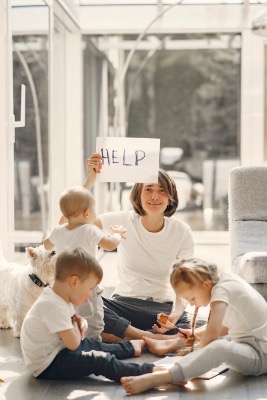 White mom in a white t-shirt sits on the floor surrounded by her three children. The children also wear white t-shirts. She holds up a sign that says “help.”