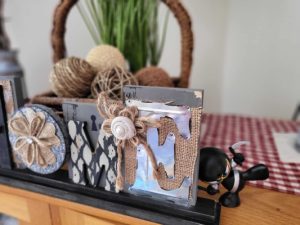 Home decor basket with green plant and linen balls is behind a rustic style HOME sign. To the right there is a ninja toy.