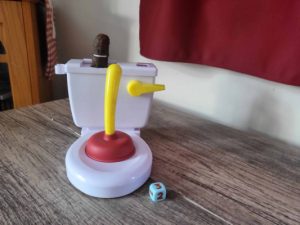 Small white toy toilet with plunger placed on it. Smiling poop log on top and a green dice next to it.