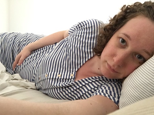 A white woman wearing a blue and white striped dress lays in bed. Her hand rests on her pregnant belly.