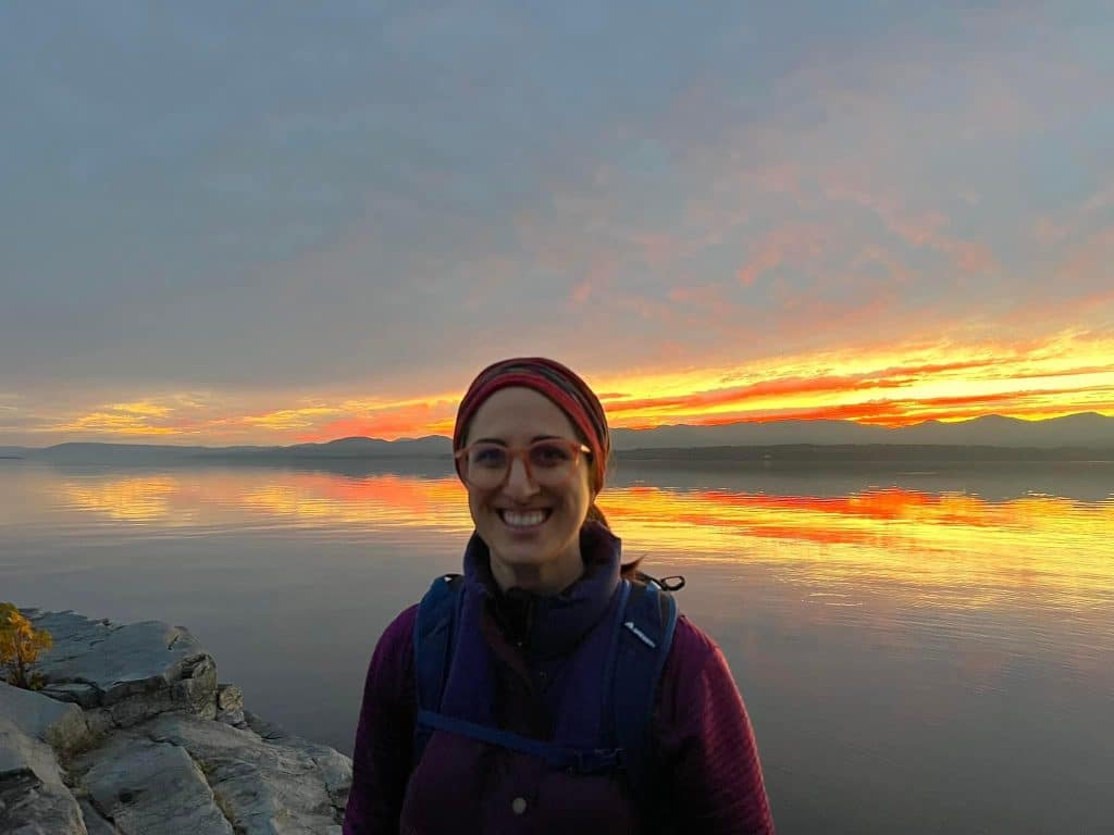 Davida standing next to a lake during the end of a sunset with orange skies.