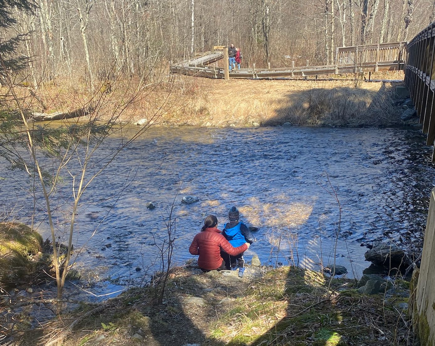 A view from behind. Davida is sitting next to a creek with her young son, holding him, as they look into the water.