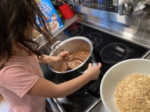 Child standing on a chair at the stove stirring chocolate and peanut butter in a pot, getting ready to add crisp rice cereal.