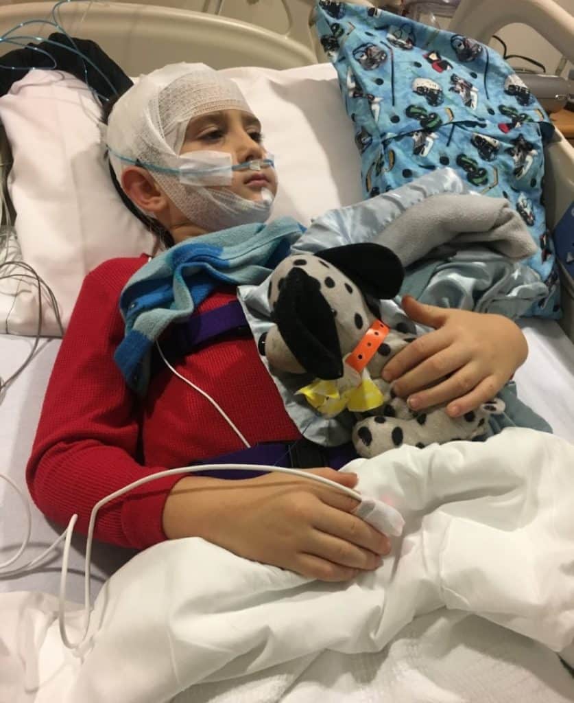 Young child wearing a red shirt and holding a stuffed animal while in a hospital bed wearing lots of gear for a sleep study to assess his pediatric sleep apnea