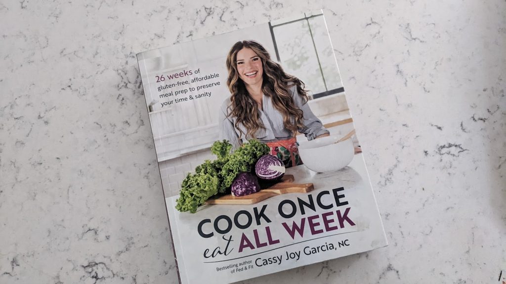 cookbook on marble countertop featuring a woman with vegetables in front of her