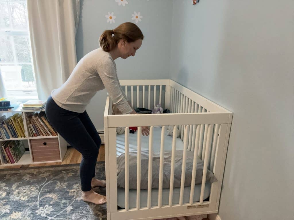 mother bends over a crib, demonstrating a correct posture to prevent back pain and injury while lifting the baby