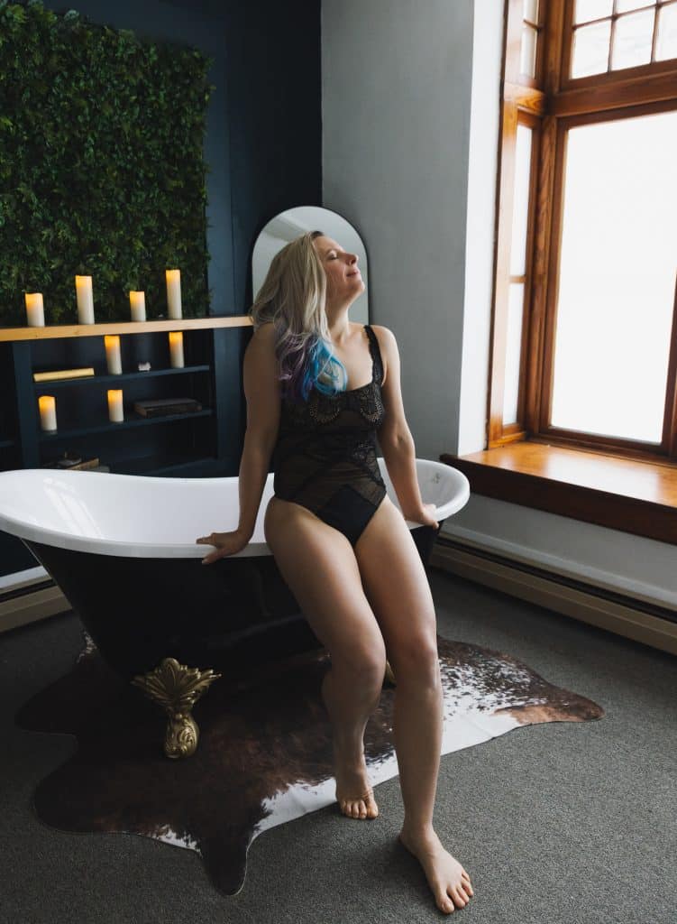 woman with blond hair with rainbow colored ends poses in front of a bathtub in lingerie.