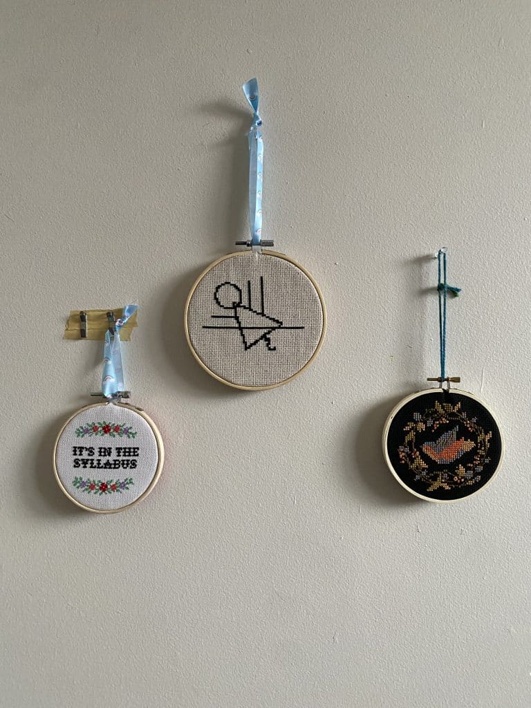 Three cross stitch projects as dead mom rituals framed in the hoop. 1: It's in the syllabus, 2: a symbol from the show Yellowjackets, 3: a bird surrounded by leaves.