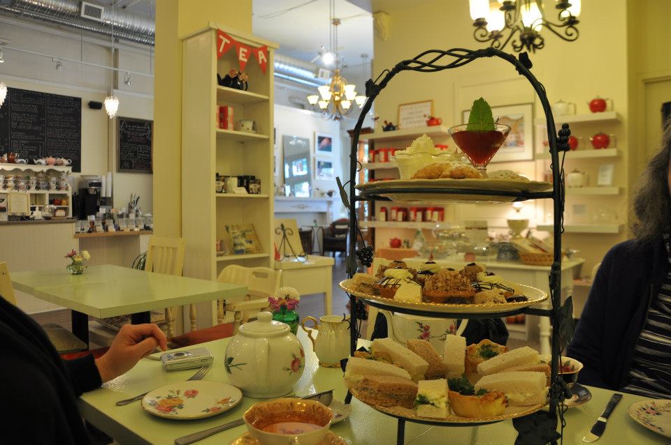 A table with a tea pot, cups and saucers, and a three-tired tray of finger sandwiches and sweets.