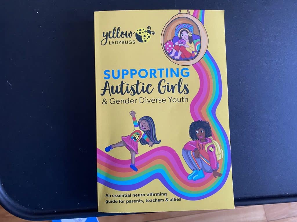 The book "Supporting autistic girls and gender diverse youth." Yellow cover with illustrations of a rainbow and three children.