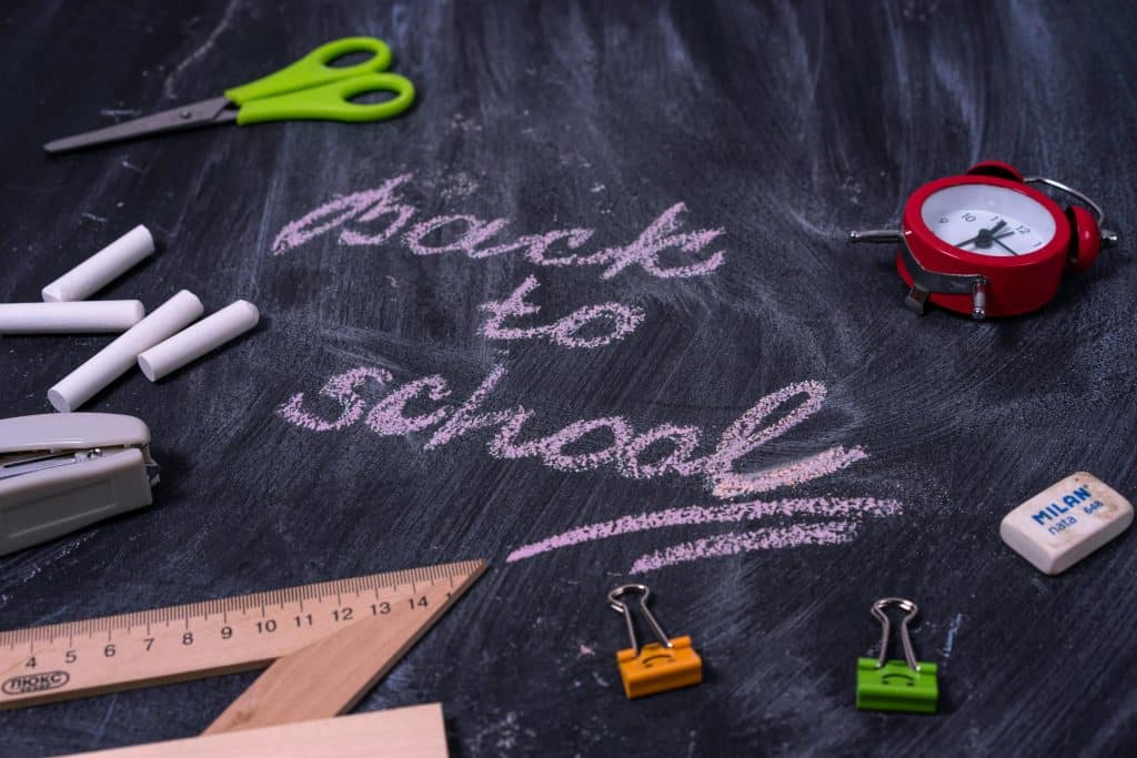 "Back to School" written on a blackboard with chalk and school supplies scattered around. 