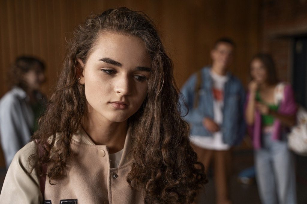 picture of a young person with long hair looking sad; behind them is a group of other young people appearing to snicker and stare.
