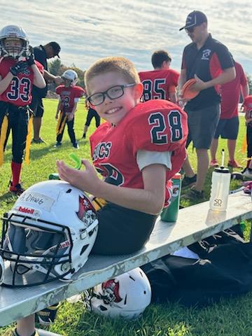 Flag football player sitting on the bench with his helmet, mouth guard, and water bottles. He is wearing black glasses and smiling.
