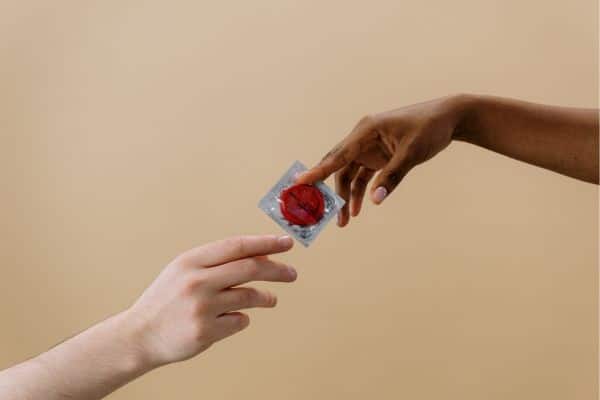 one hand handing another a condom against a tan background. male birth control.