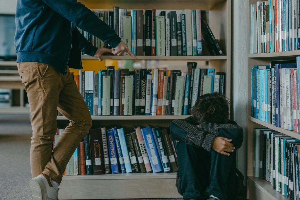 person pointing at a cowering figure on the floor of a library in a bullying pose.