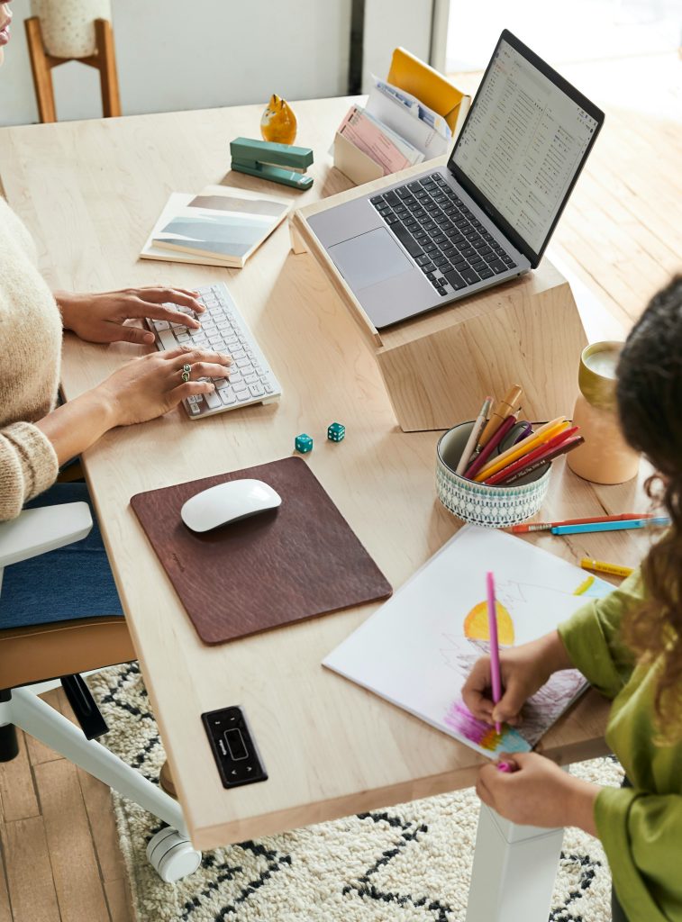 modern motherhood: a mom types on the computer as her child colors on the desk next to her.